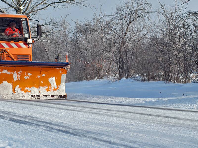 Ein orangefarbener Schneepflug fährt auf einer schneebedeckten Straße zwischen winterlichen Bäumen.