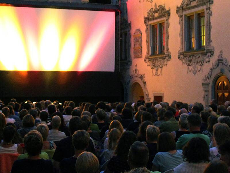 Eine Menschenmenge sitzt abends im Freien vor einer großen Leinwand zwischen historischen Gebäuden beim Ansehen eines Films.