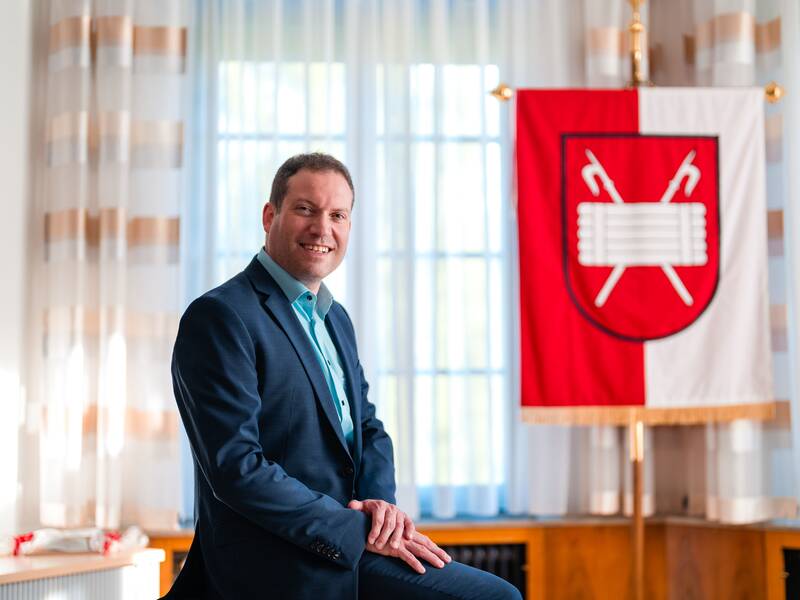 Bürgermeister Frank Zimmermann in blauem Anzug, in seinem Büro, sitzend auf seinem Schreibtisch, im Hintergrund die Gaildorfer Stadtfahne