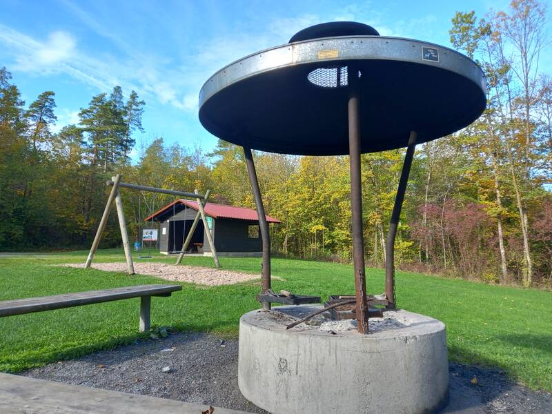 Spiel- und Grillplatz Kirgel, im Vordergrund eine Grillstelle mit Sitzbank, im Hintergrund ein mit rotem Dach versehene Schutzhütte