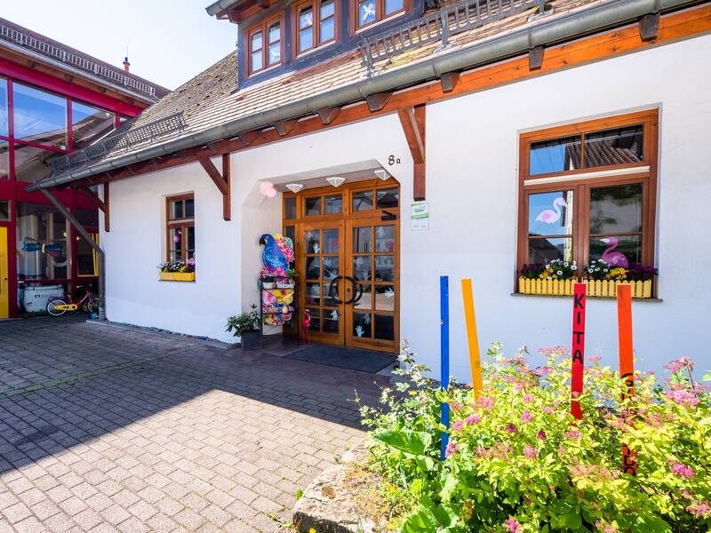 KIndergarten Großaltdorf, Eingang mit bunten Holzstäben, roter Tür und im Hintergrund erkennbarer Anbau