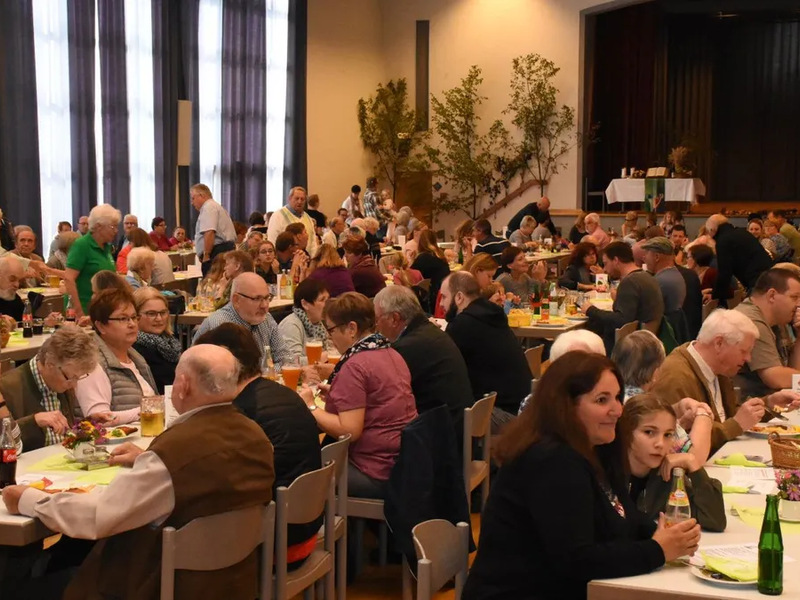 Viele Menschen an weißen Tischen sitzend in einer geschmückten Halle mit Getränken und Essen vor sich