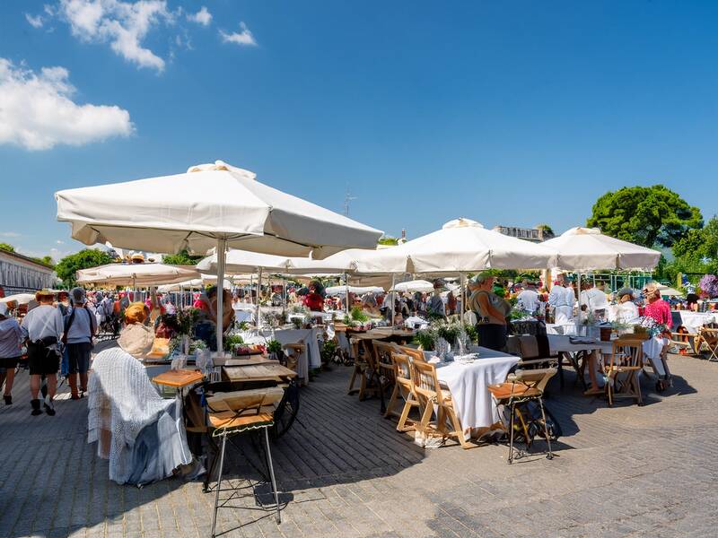 KI generierter Flohmarkt mit vielen Menschen und Tischen auf einer Straße unter freiem Himmel mit Sonnenschirmen