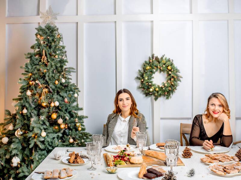 KI 2 Menschen sitzen an Tischen mit weihnachtlichem Gebäck, Rednerpult im Hintergrund, geschmückter Weihnachtsbaum im Vordergrund