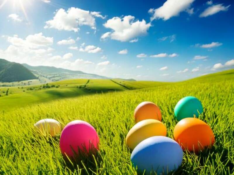 Symbolbild Ostern Bunte Ostereier auf einer grünen Wiese, im Hintergrund blauer Himmel mit weißen Wolken und einer strahlenden Sonne