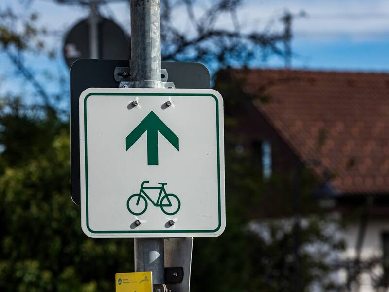 Ein weißes Schild mit einem grünen Fahrrad und Richtungspfeil als Wegweiser für einen Radweg.  Das Schild ist an einem Laternenpfosten befestigt.